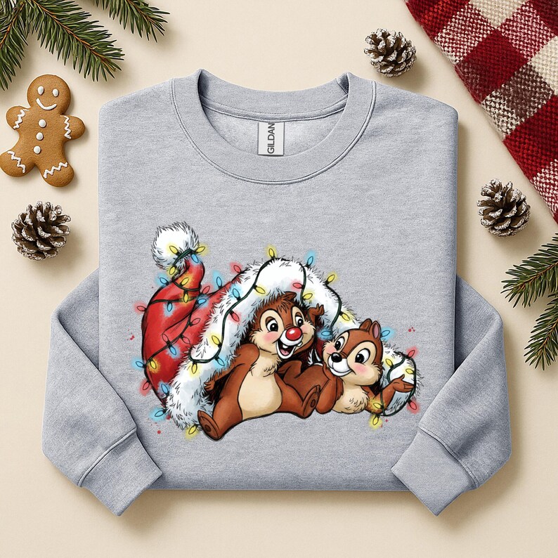 May include: A light gray sweatshirt featuring a festive graphic of two cartoon chipmunks nestled under a Santa hat adorned with colorful Christmas lights. The design is set against a backdrop of holiday decorations.