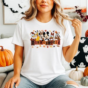 May include: White t-shirt featuring Disney characters: Donald Duck, Minnie Mouse, Mickey Mouse, Daisy Duck, and Goofy. The word "Disneyworld" is printed in brown letters. Autumn leaves and pumpkins are in the background.