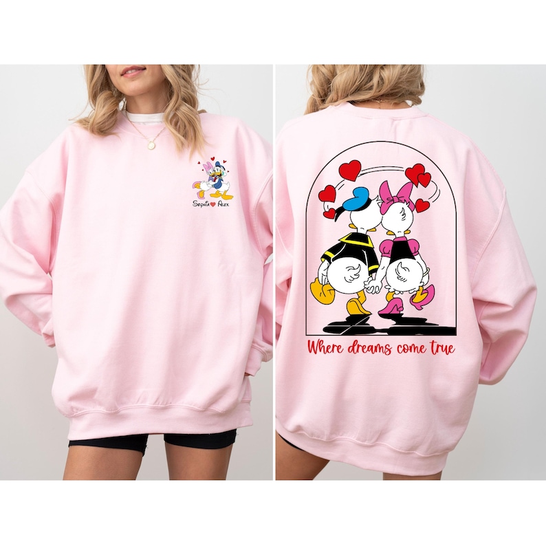 May include: Pink sweatshirt with a cartoon image of Donald and Daisy Duck on the back. The text "Where dreams come true" is printed below the image. The front of the sweatshirt has a smaller image of Donald and Daisy Duck with the text "Say it with a Duck".
