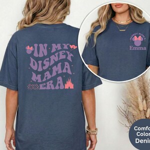 Puede incluir: Camisetas azul denim con gr&aacute;ficos tem&aacute;ticos de Disney. Una camiseta presenta "IN MY DISNEY MAMA ERA" en morado y rosa. La otra camiseta tiene "DISNEY MAMA" con un lazo y el nombre "Emma".