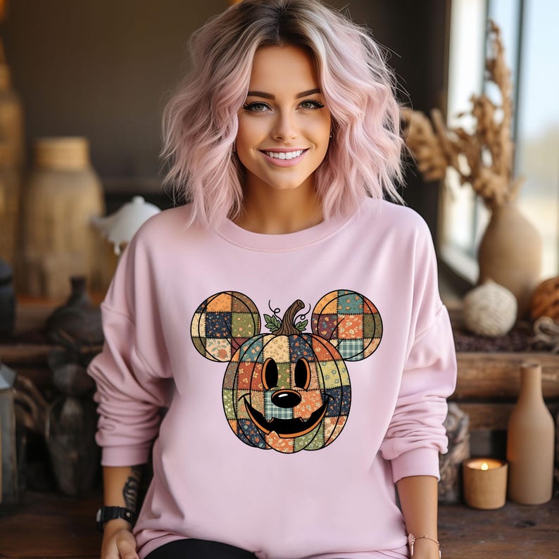 May include: A light pink sweatshirt featuring a patchwork pumpkin design with Mickey Mouse ears. The pumpkin has a smiling face and is made of various fabric patterns in fall colors. The sweatshirt has long sleeves.