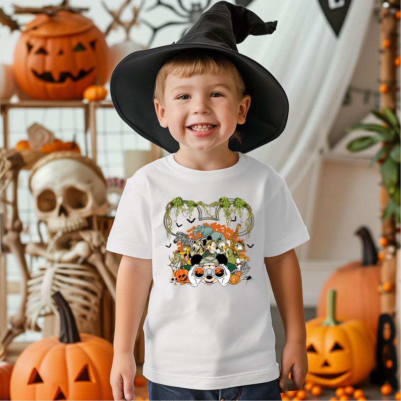 May include: A young child wearing a black witch hat and a white t-shirt with a Halloween-themed graphic. The graphic features cartoon characters, pumpkins, and bats. The background includes Halloween decorations like pumpkins and a skeleton.