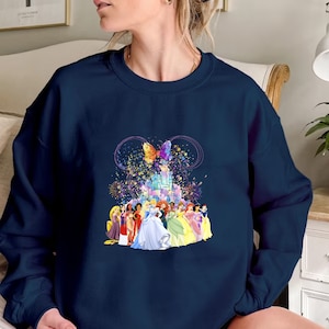Puede incluir: Una sudadera azul marino con un gr&aacute;fico colorido de princesas Disney frente al castillo de Cenicienta. El gr&aacute;fico tambi&eacute;n incluye una mariposa y brillos.