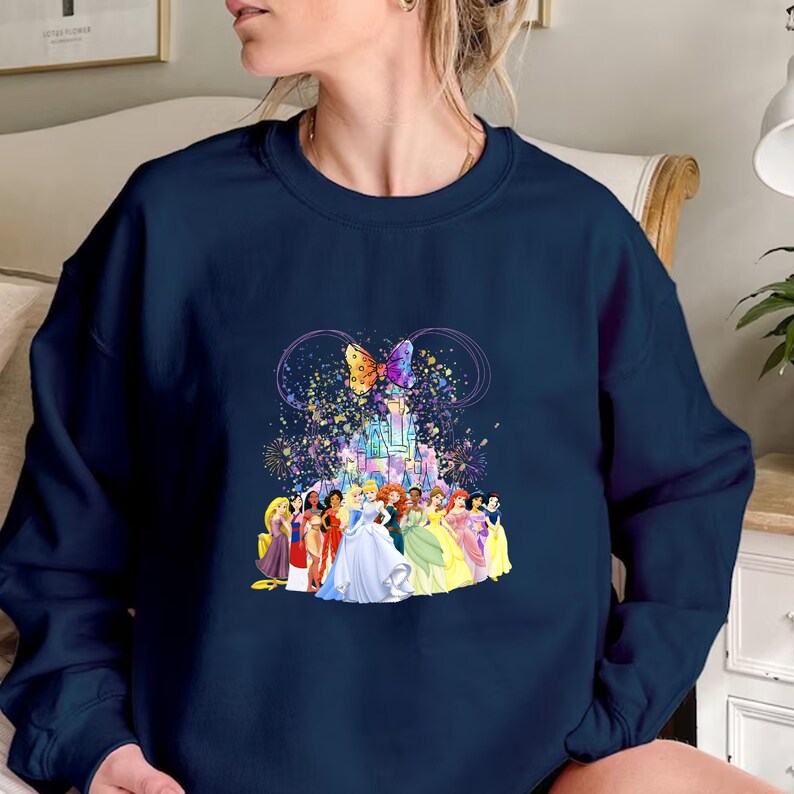 Puede incluir: Una sudadera azul marino con un gr&aacute;fico colorido de princesas Disney frente al castillo de Cenicienta. El gr&aacute;fico tambi&eacute;n incluye una mariposa y brillos.
