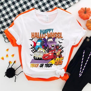 Może przedstawiać: Biała koszulka z grafiką kresk&oacute;wek samochod&oacute;w przebranych na Halloween. Na koszulce nadrukowany jest tekst "Happy Hallo-Wheel Truck or Treat".