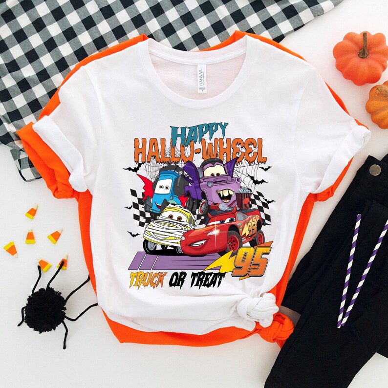 Może przedstawiać: Biała koszulka z grafiką kresk&oacute;wek samochod&oacute;w przebranych na Halloween. Na koszulce nadrukowany jest tekst "Happy Hallo-Wheel Truck or Treat".