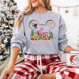 May include: A heather gray sweatshirt featuring a colorful graphic of Disney characters and a castle, with the word "Christmas" below. The design is framed by Mickey Mouse ears. The person is wearing red and white plaid pajama pants.