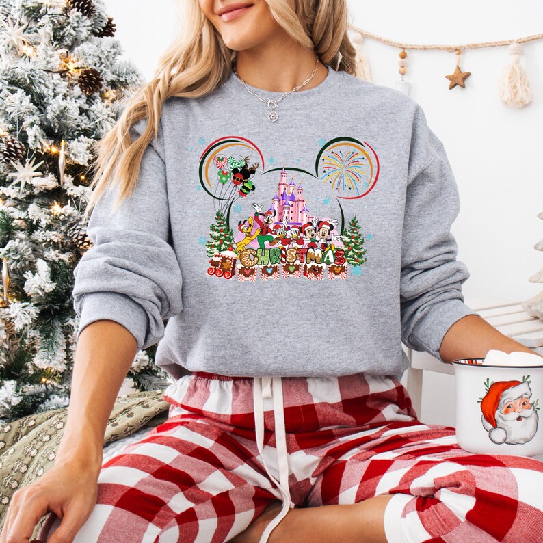 May include: A heather gray sweatshirt featuring a colorful graphic of Disney characters and a castle, with the word "Christmas" below. The design is framed by Mickey Mouse ears. The person is wearing red and white plaid pajama pants.