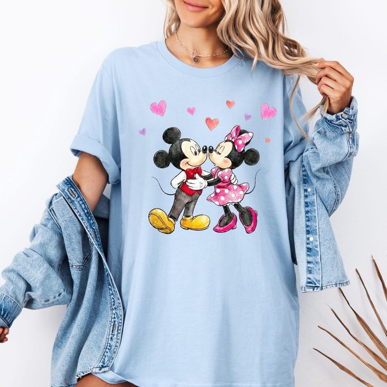 May include: Pale blue t-shirt with a watercolour illustration of Mickey and Minnie Mouse kissing. Mickey is in a red shirt, grey trousers, and yellow shoes. Minnie wears a pink polka dot dress and shoes. Pink hearts are scattered around.