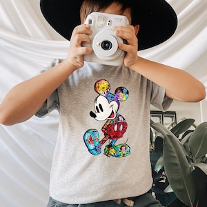 Puede incluir: Una camiseta gris con un gr&aacute;fico de Mickey Mouse colorido. El gr&aacute;fico presenta a Mickey Mouse con un collage de personajes de Disney en su cuerpo.