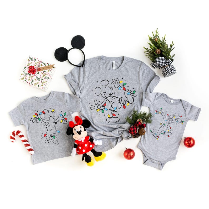 May include: Gray t-shirts and a onesie featuring Mickey and Minnie Mouse with Christmas lights. A Minnie Mouse plush, Mickey Mouse ears, and holiday decorations are also present. Perfect for a Disney-themed holiday.