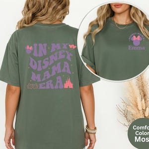 Puede incluir: Camiseta verde musgo con el texto "IN MY DISNEY MAMA ERA" en morado y rosa. La parte delantera de la camiseta tiene un peque&ntilde;o dise&ntilde;o con el texto "DISNEY MAMA" y un lazo, con el nombre "Emma" debajo.