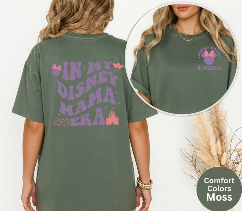 Puede incluir: Camiseta verde musgo con el texto "IN MY DISNEY MAMA ERA" en morado y rosa. La parte delantera de la camiseta tiene un peque&ntilde;o dise&ntilde;o con el texto "DISNEY MAMA" y un lazo, con el nombre "Emma" debajo.