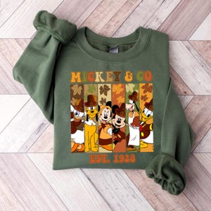May include: Green sweatshirt featuring the text "MICKEY & CO" and "EST. 1928". The design includes cartoon characters like Mickey Mouse, Donald Duck, and Goofy, all wearing pilgrim hats and fall-themed attire. The sweatshirt is a fall-themed item.