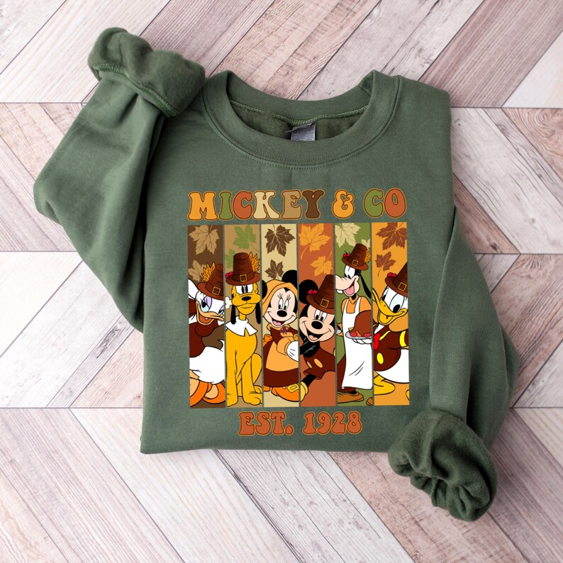 May include: Green sweatshirt featuring the text "MICKEY & CO" and "EST. 1928". The design includes cartoon characters like Mickey Mouse, Donald Duck, and Goofy, all wearing pilgrim hats and fall-themed attire. The sweatshirt is a fall-themed item.
