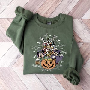 May include: Green sweatshirt featuring a Halloween-themed graphic with Disney characters like Mickey Mouse, Minnie Mouse, Goofy, Donald Duck, and Pluto. The design includes a carved pumpkin and bats.