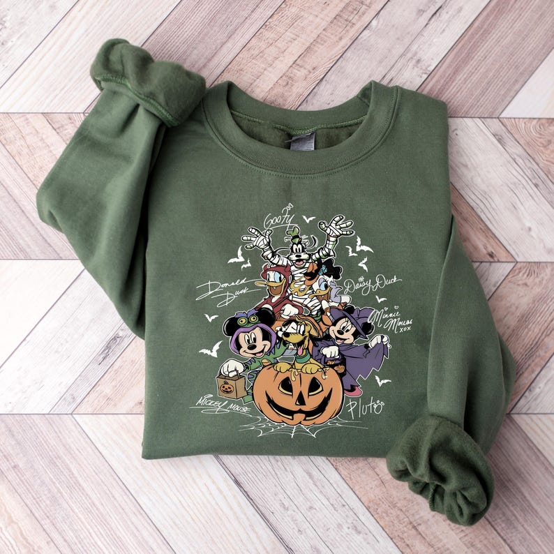 May include: Green sweatshirt featuring a Halloween-themed graphic with Disney characters like Mickey Mouse, Minnie Mouse, Goofy, Donald Duck, and Pluto. The design includes a carved pumpkin and bats.