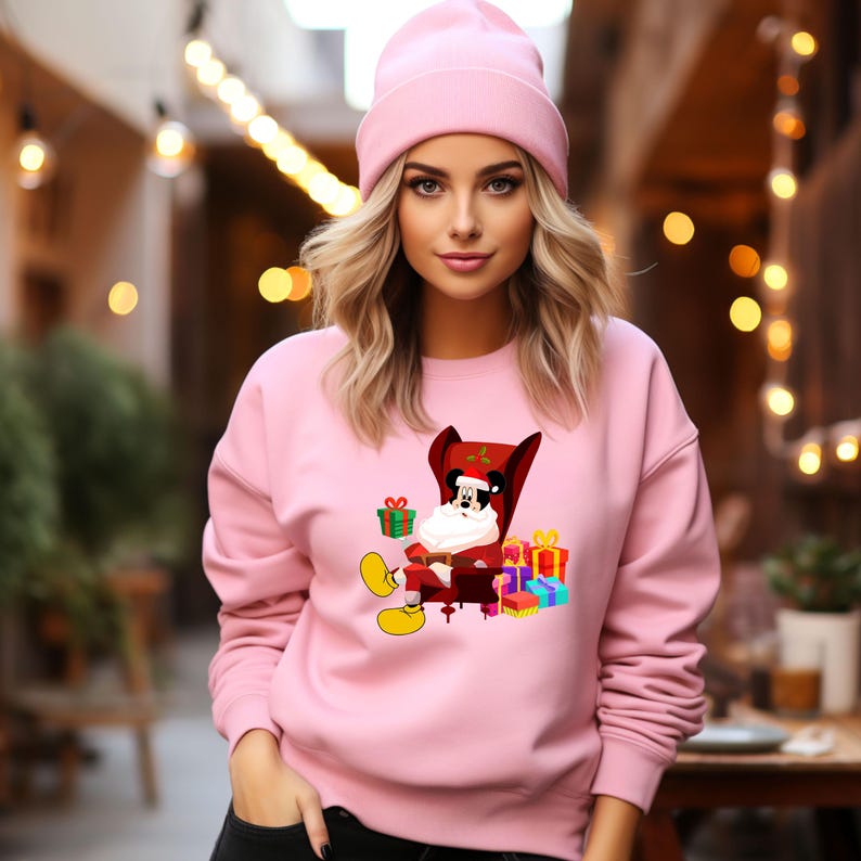 Op de afbeelding: Een lichtroze sweatshirt en beanie. Het sweatshirt heeft een cartoon van Mickey Mouse verkleed als kerstman, zittend in een rode stoel met cadeaus. De beanie past bij de kleur van het sweatshirt. De foto is buiten genomen.