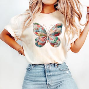 May include: Cream-colored t-shirt featuring a large, colorful butterfly design. The butterfly is made up of various fabric patches in shades of red, pink, green, and blue, giving it a quilted appearance. The shirt is paired with blue denim jeans.