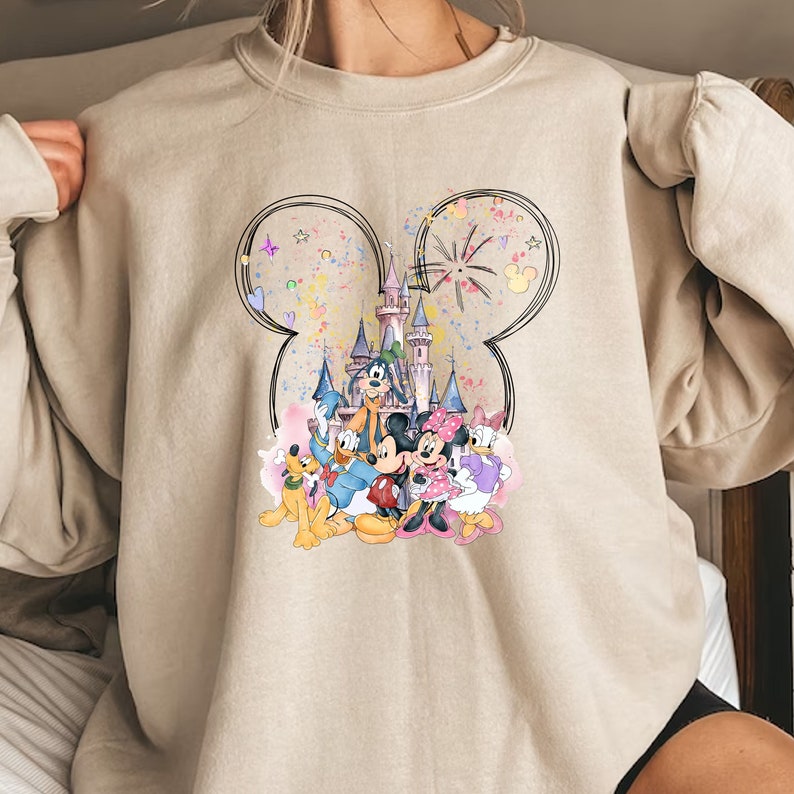 May include: A beige sweatshirt with a colorful graphic print of Mickey Mouse, Minnie Mouse, Donald Duck, Goofy, and Pluto in front of Cinderella's Castle. The graphic is surrounded by a black outline of Mickey Mouse's ears.