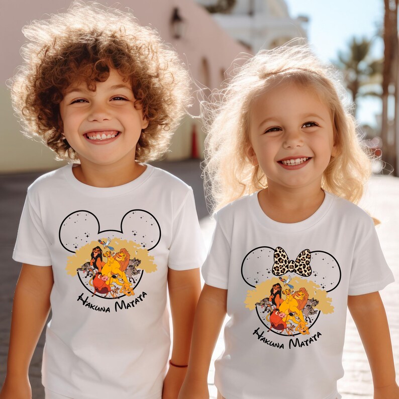 Puede incluir: Dos ni&ntilde;os con camisetas blancas con un dise&ntilde;o de Disney El Rey Le&oacute;n. Las camisetas presentan una silueta de oreja de Mickey con los personajes de El Rey Le&oacute;n en su interior. El texto "Hakuna Matata" est&aacute; impreso debajo del dise&ntilde;o. Una camiseta tiene un lazo en la oreja de Mickey y la otra tiene una oreja de Mickey normal.