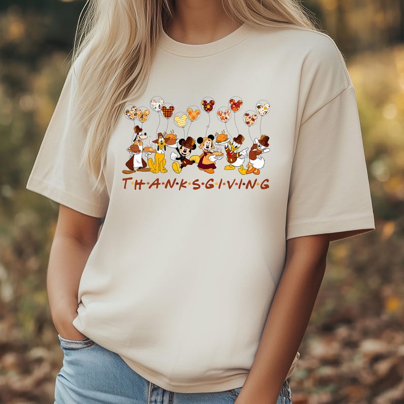 Pu&ograve; includere: T-shirt beige con grafica a tema Ringraziamento. Il design include personaggi Disney che tengono cibo e palloncini. La parola "THANKSGIVING" &egrave; stampata in lettere marroni nella parte inferiore della maglietta.