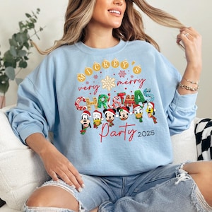 K&ouml;nnte beinhalten: Hellblauer Pullover mit einem Mickey Mouse Weihnachtsparty-Design. Das Design zeigt Mickey Mouse und seine Freunde, darunter Donald Duck, Goofy und Pluto. Der Text auf dem Pullover lautet "Mickey's very merry Christmas Party 2025".