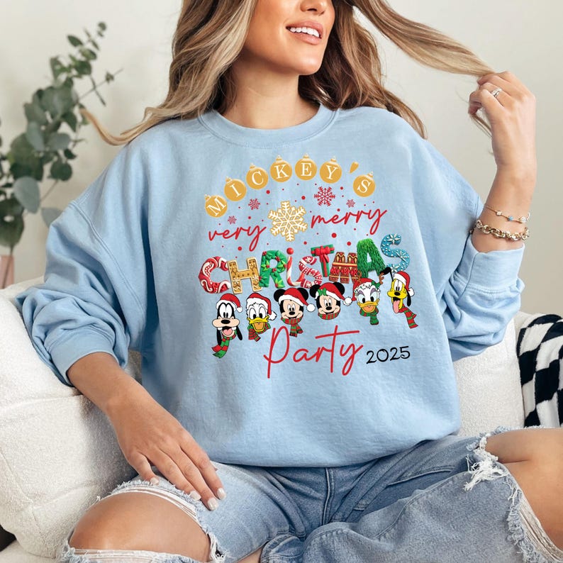 K&ouml;nnte beinhalten: Hellblauer Pullover mit einem Mickey Mouse Weihnachtsparty-Design. Das Design zeigt Mickey Mouse und seine Freunde, darunter Donald Duck, Goofy und Pluto. Der Text auf dem Pullover lautet "Mickey's very merry Christmas Party 2025".