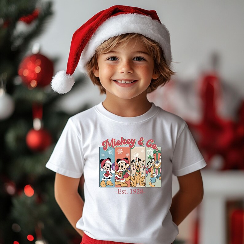 May include: A child wearing a white t-shirt with a colorful graphic of Mickey Mouse and friends in holiday attire. The shirt reads "Mickey & Co - Est. 1928." The child is also wearing a red and white Santa hat, smiling in front of a Christmas tree.