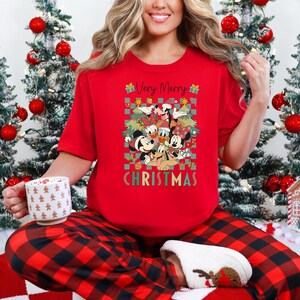 May include: A red Christmas t-shirt featuring Mickey Mouse, Minnie Mouse, Donald Duck, and Goofy. The shirt has the text "Very Merry Christmas" in green and white. The person is wearing red and black plaid pajama pants and holding a white mug.