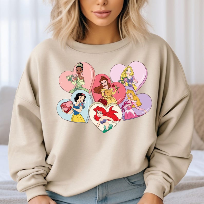 Pode incluir: Uma camisa de moletom bege com uma estampa gr&aacute;fica de princesas Disney em cora&ccedil;&otilde;es. As princesas s&atilde;o Branca de Neve, Cinderela, Ariel, Bela, Tiana e Rapunzel.