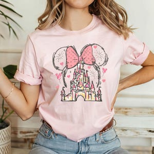 May include: Pink t-shirt with a graphic of a pink bow, fireworks, and a pink castle. The castle is a stylized version of Cinderella's Castle.