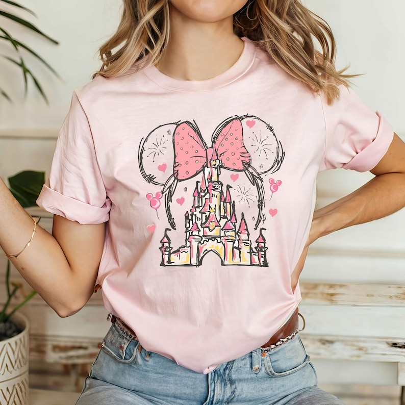 May include: Pink t-shirt with a graphic of a pink bow, fireworks, and a pink castle. The castle is a stylized version of Cinderella's Castle.