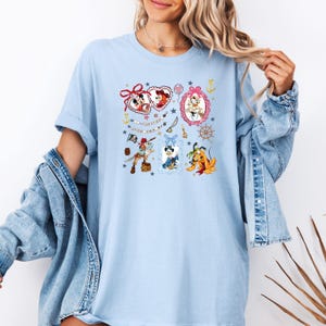 May include: Light blue t-shirt with a colorful graphic print featuring cartoon characters in frames. The characters include Mickey Mouse, Minnie Mouse, Pluto, and Donald Duck. The text on the shirt reads "It's a Small World".