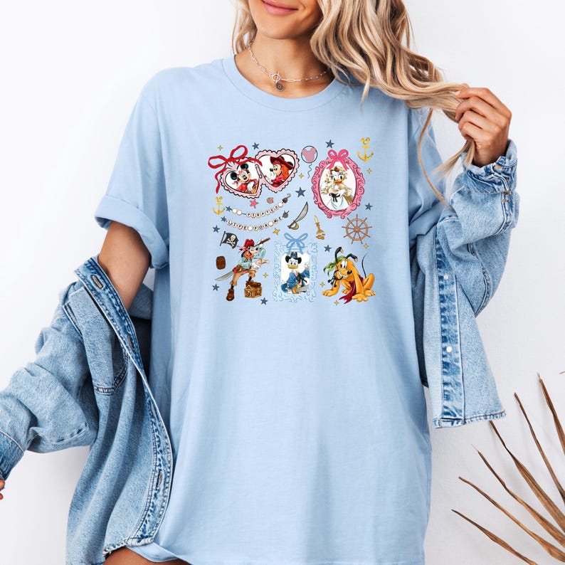 May include: Light blue t-shirt with a colorful graphic print featuring cartoon characters in frames. The characters include Mickey Mouse, Minnie Mouse, Pluto, and Donald Duck. The text on the shirt reads "It's a Small World".
