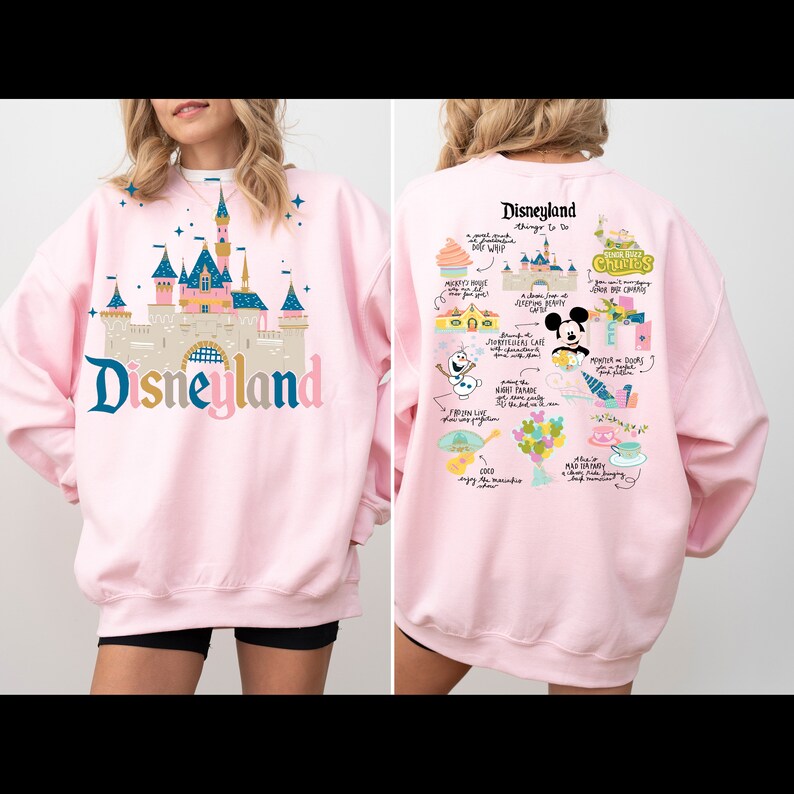 May include: Pink sweatshirt with a graphic print of Disneyland's Sleeping Beauty Castle on the front and a map of Disneyland attractions on the back. The text "Disneyland" is printed in large letters on the front of the sweatshirt.