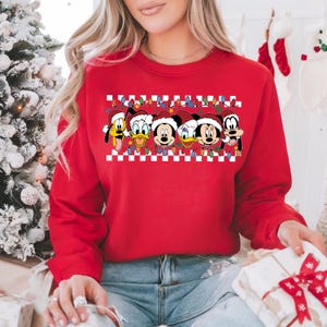 May include: Red Christmas sweater with a checkered pattern and a row of Disney characters wearing Santa hats. The characters include Mickey Mouse, Minnie Mouse, Donald Duck, Goofy, and Pluto.