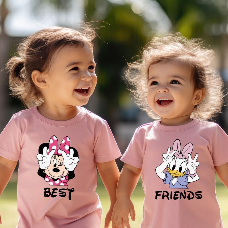 Może przedstawiać: Dwie małe dziewczynki w r&oacute;żowych koszulkach z postaciami z kresk&oacute;wek. Dziewczynka po lewej stronie ma wz&oacute;r Minnie Mouse z napisem "BEST", a dziewczynka po prawej stronie ma wz&oacute;r Daisy Duck z napisem "FRIENDS".