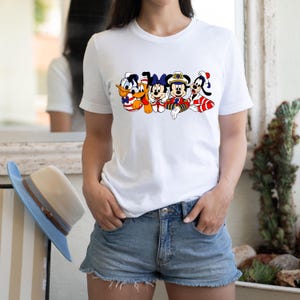 Peut inclure: T-shirt blanc avec un motif de Mickey Mouse, Donald Duck, Dingo et Pluto portant des tenues de marin. Les personnages sourient et saluent.