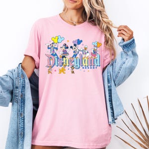 May include: Pink t-shirt featuring a graphic of Mickey Mouse, Minnie Mouse, Donald Duck, Goofy, and Pluto in front of a Disneyland Resort logo. The characters are holding balloons and stars.