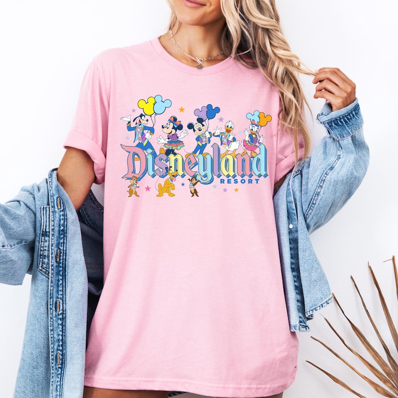 May include: Pink t-shirt featuring a graphic of Mickey Mouse, Minnie Mouse, Donald Duck, Goofy, and Pluto in front of a Disneyland Resort logo. The characters are holding balloons and stars.