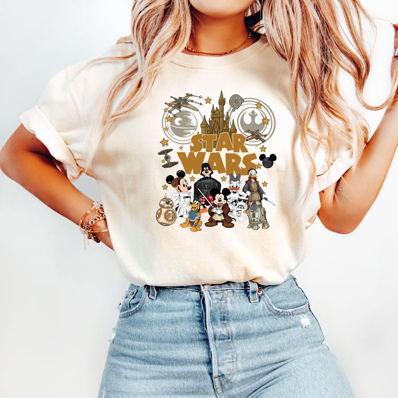May include: Cream-colored t-shirt featuring a graphic design with the words "STAR WARS" and various characters from the Star Wars and Disney universes. The design includes Mickey Mouse, Darth Vader, and R2-D2.