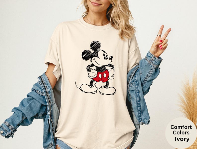 May include: Ivory-colored t-shirt featuring a black and red Mickey Mouse graphic. The cartoon character is drawn in a classic style, with black ears, a white face, and red shorts. The shirt is part of the "Comfort Colors" collection.
