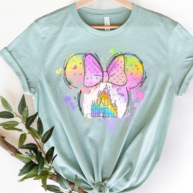 Puede incluir: Una camiseta azul claro con un gr&aacute;fico colorido de orejas de Mickey Mouse con un lazo rosa y un castillo de colores arco&iacute;ris en su interior. El gr&aacute;fico tiene un efecto acuarela.