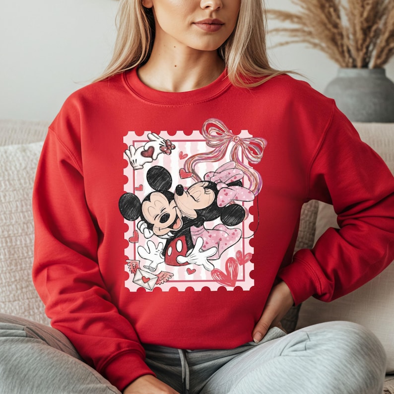 May include: A red sweatshirt featuring a graphic of Mickey and Minnie Mouse embracing within a postage stamp design. The stamp includes hearts, a bow, and a love letter. The sweatshirt is a solid red color.