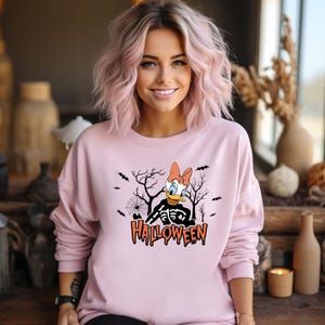 May include: A light pink sweatshirt with a Halloween-themed graphic. The graphic features a cartoon character with a skeleton body, bats, a spiderweb, and the word "HALLOWEEN" in orange. The sweatshirt has long sleeves.