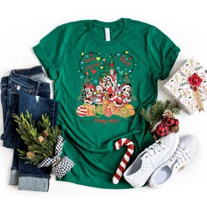 May include: Green Christmas-themed t-shirt with a graphic featuring Disney characters in Santa outfits, a castle, and the words "Very Merry Christmas Party 2023." The shirt is paired with jeans, sneakers, and a wrapped gift.