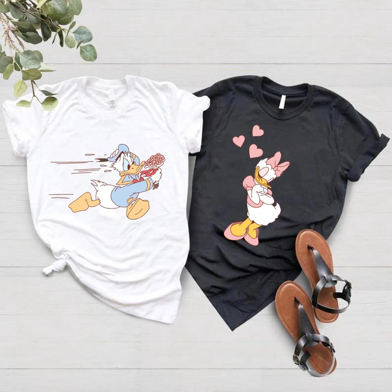May include: A white t-shirt with a cartoon image of Donald Duck running with a bouquet of flowers. A black t-shirt with a cartoon image of Daisy Duck with hearts floating above her head.