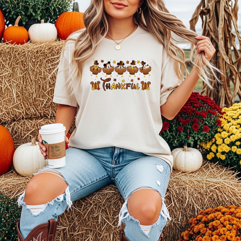 May include: Beige t-shirt with a Thanksgiving-themed graphic. The graphic features cartoon turkeys wearing pilgrim hats and the text "Thankful". The shirt is paired with blue jeans. Pumpkins and hay bales are in the background.