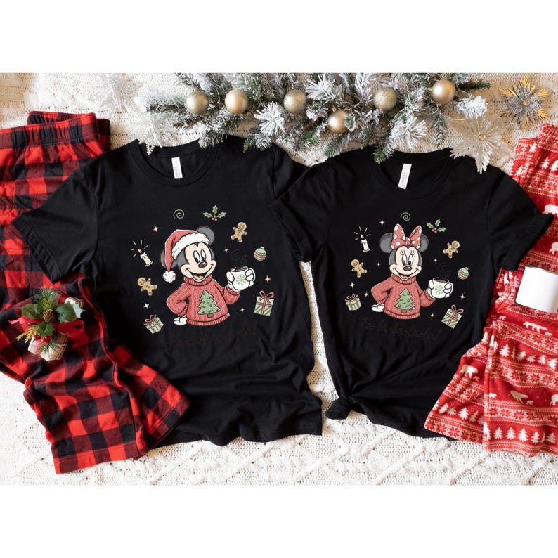 May include: Two black t-shirts featuring Mickey and Minnie Mouse in Christmas sweaters, holding mugs. The shirts have festive graphics including gingerbread men, candles, and gift boxes. Red and black plaid pajama pants are visible on the sides.